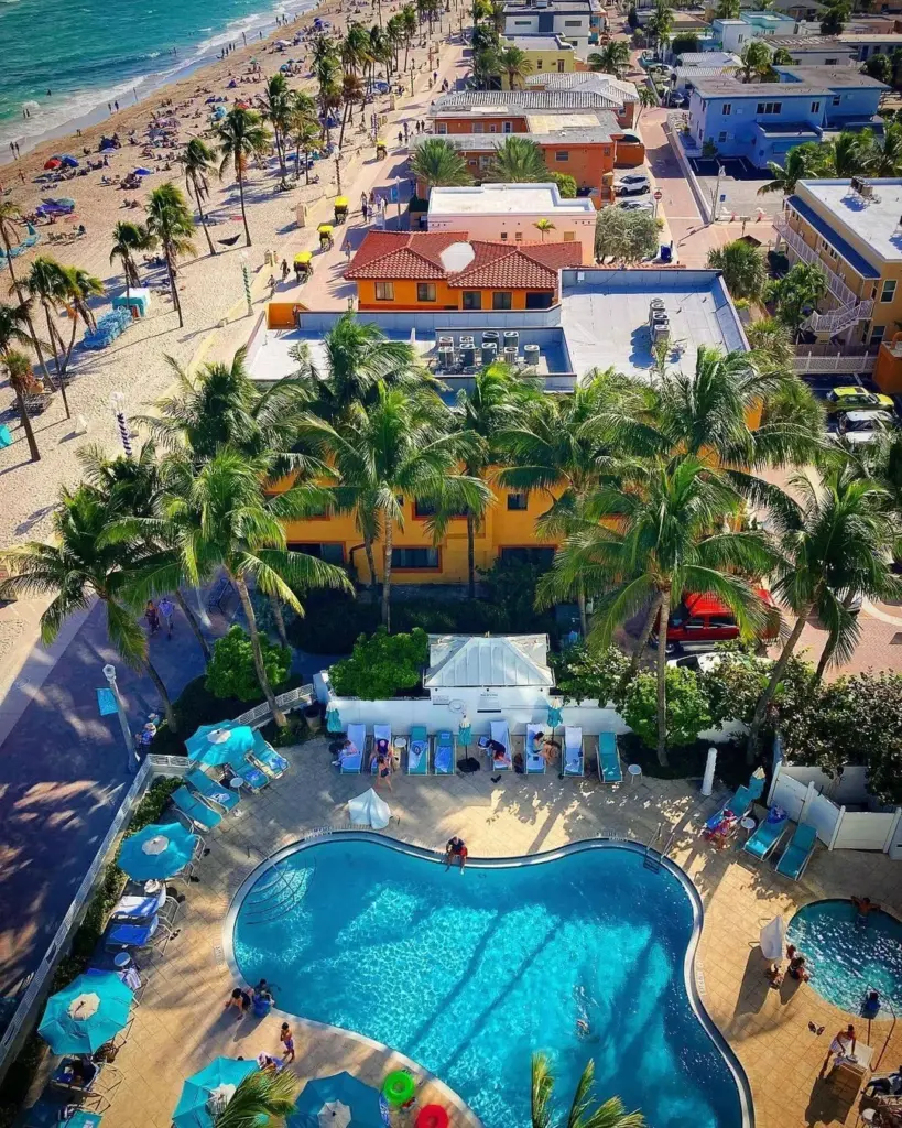 Aerial view of Hollywood beach pool area with palm trees, colorful buildings and the beach area with beach umbrellas and people. 